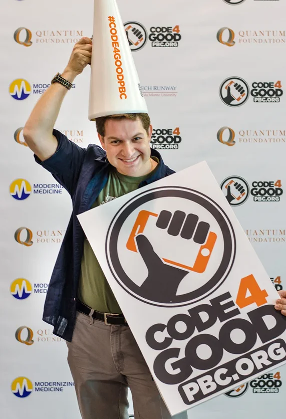 Roman Smolkin at a Code for Good event, holding a Code4Good PBC sign
                    and balancing a #CODE4GOODPBC megaphone on his head.